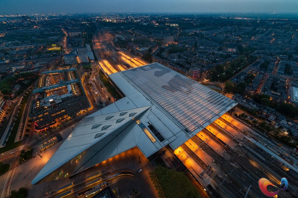 Vleugelvormig futuristisch gebouw boven druk spoor en stationsgebied bij schemering met oranje perronverlichting.
