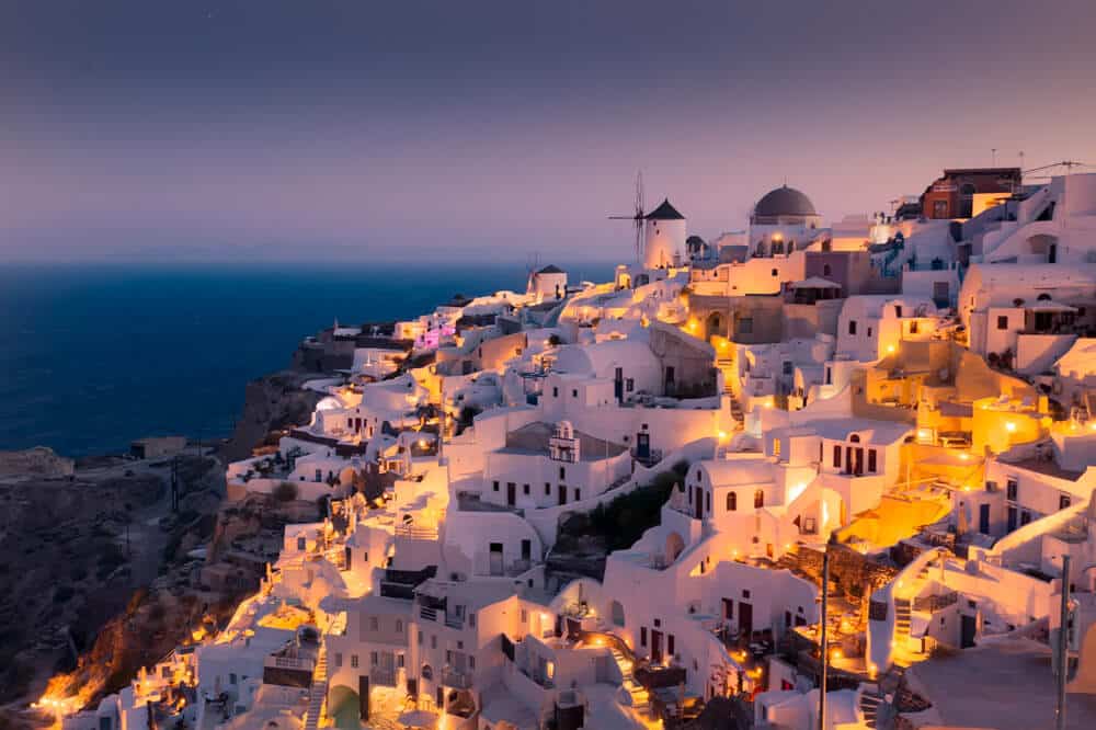 Santorini caldera dorp schemerlicht met witte kubuswoningen, koepelkerk en windmolen boven zee.