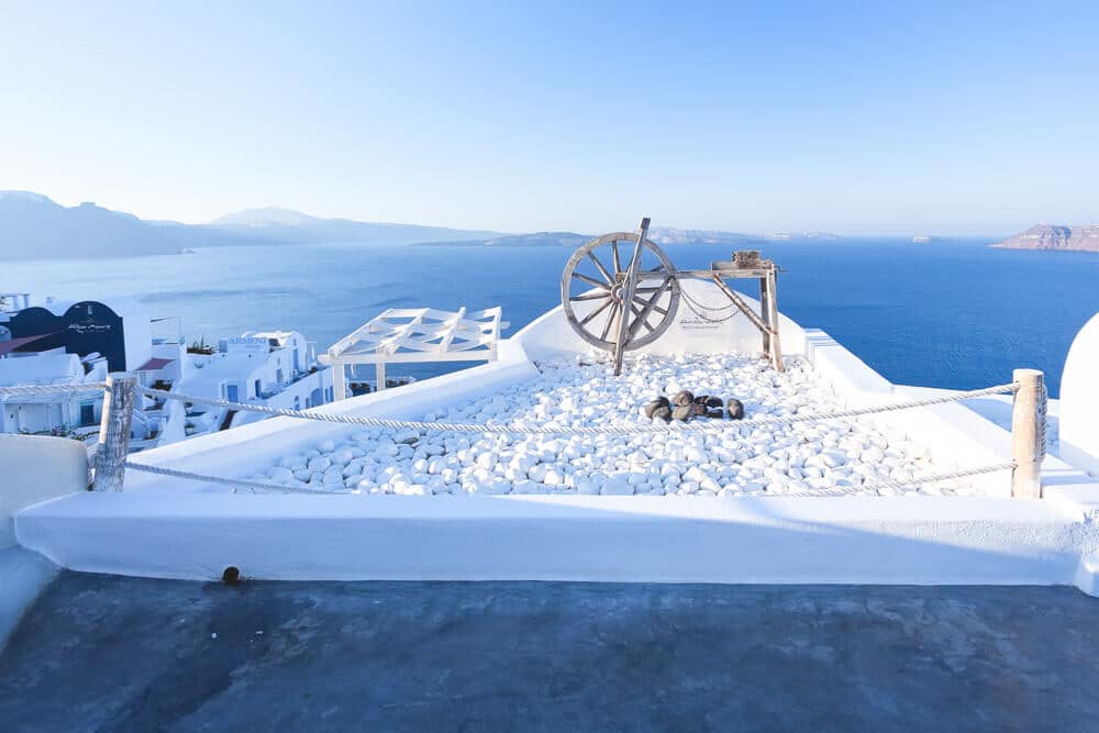 Mediterrane dakterras met witte gebouwen, wagenwiel en touwbarrière boven een azuurblauwe zee.