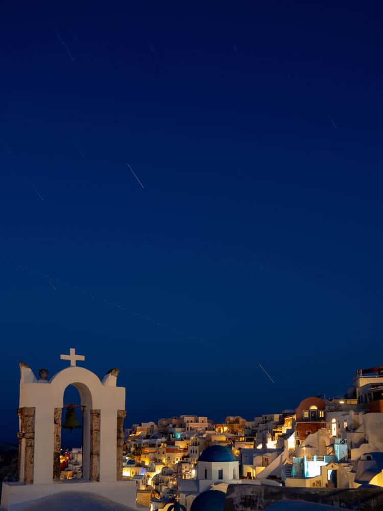 Santorini: witte kubuswoningen op terrassen, blauwe koepels, bel in boog met kruis bij sterrenhemel.