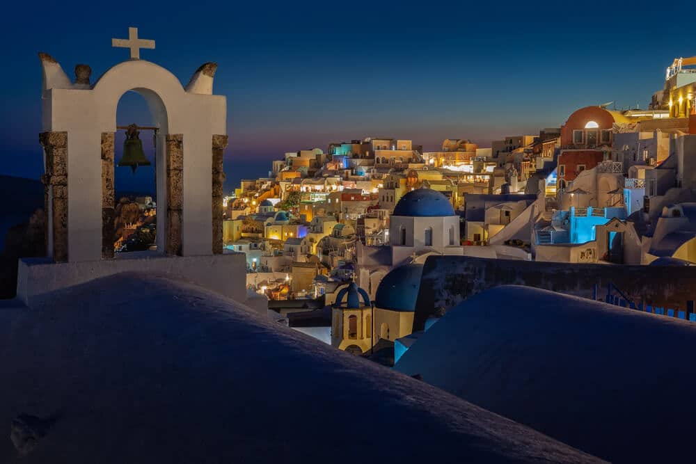 Santorini: wit gepleisterde klokkentoren en azuurblauwe koepels boven witte huizen bij schemerlicht.