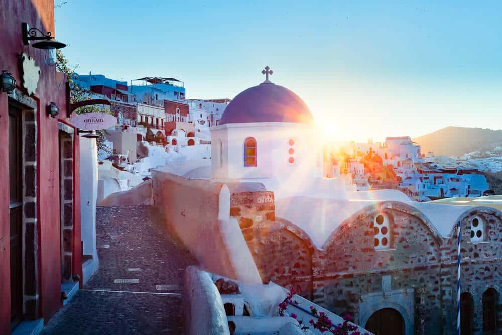 Santorini: witte kerk met diepe blauwe koepel bij gouden uur, kronkelende straat en terrassen.