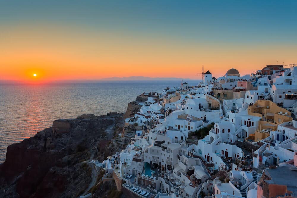 Santorini zonsondergang boven witte kubuswoningen, windmolen en blauwe deuren aan zee.