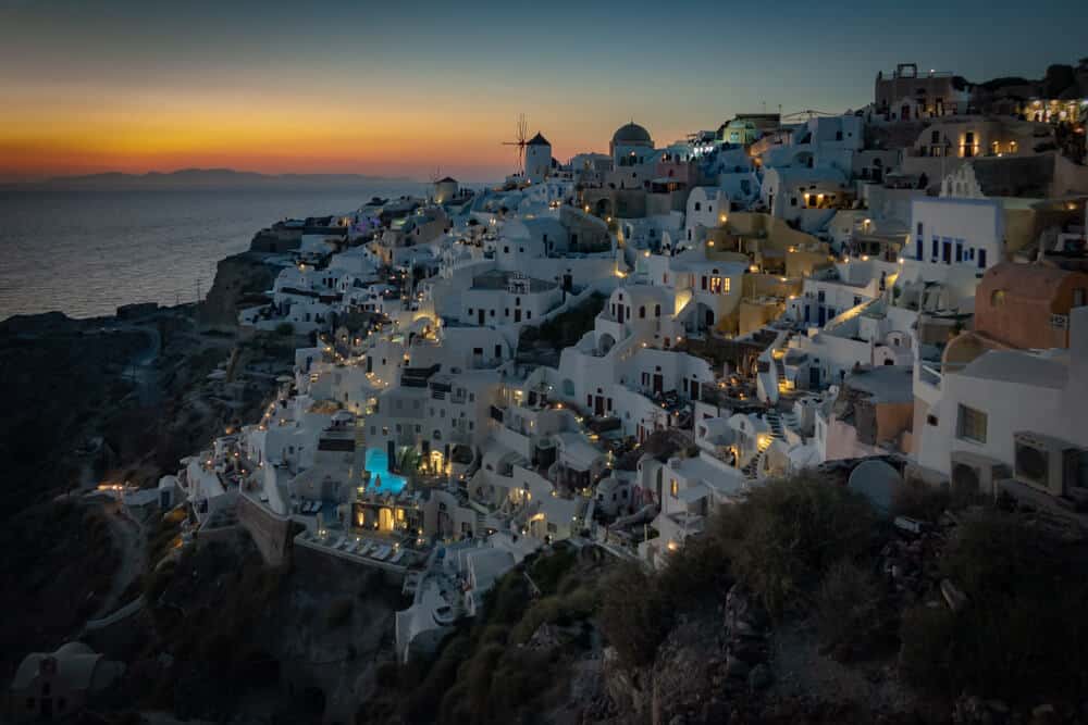 Santorini windmolen boven witte calderahuizen bij zonsondergang met blauwe deuren en turkoois zwembad.