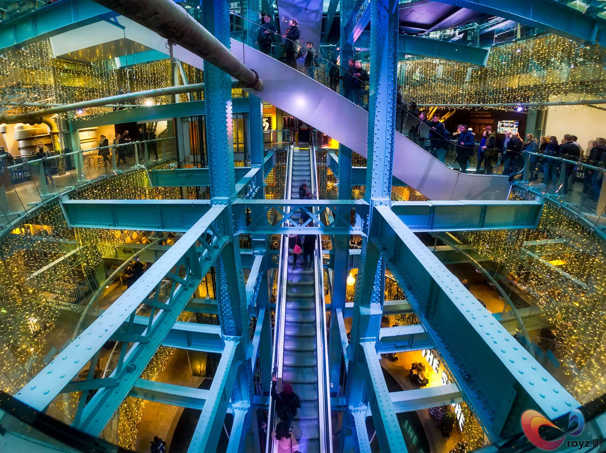 Futuristisch atrium met tealblauw staalwerk, centrale kern, glazen balustrades en roltrap; bezoekers in winterjassen.