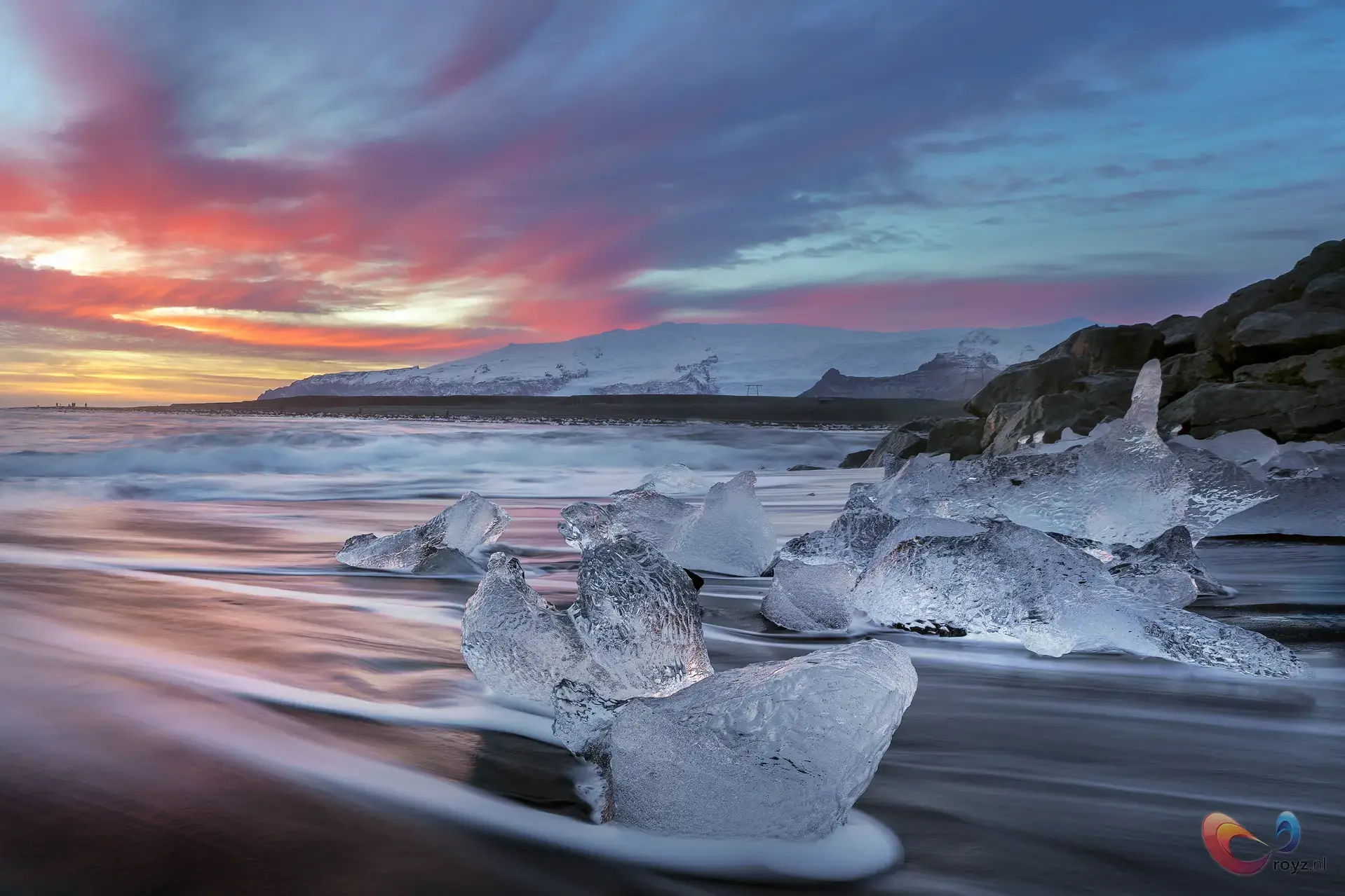 Zonsondergang op zwarte zandkust van IJsland met ijsblokken en rotsachtige uitlopers.