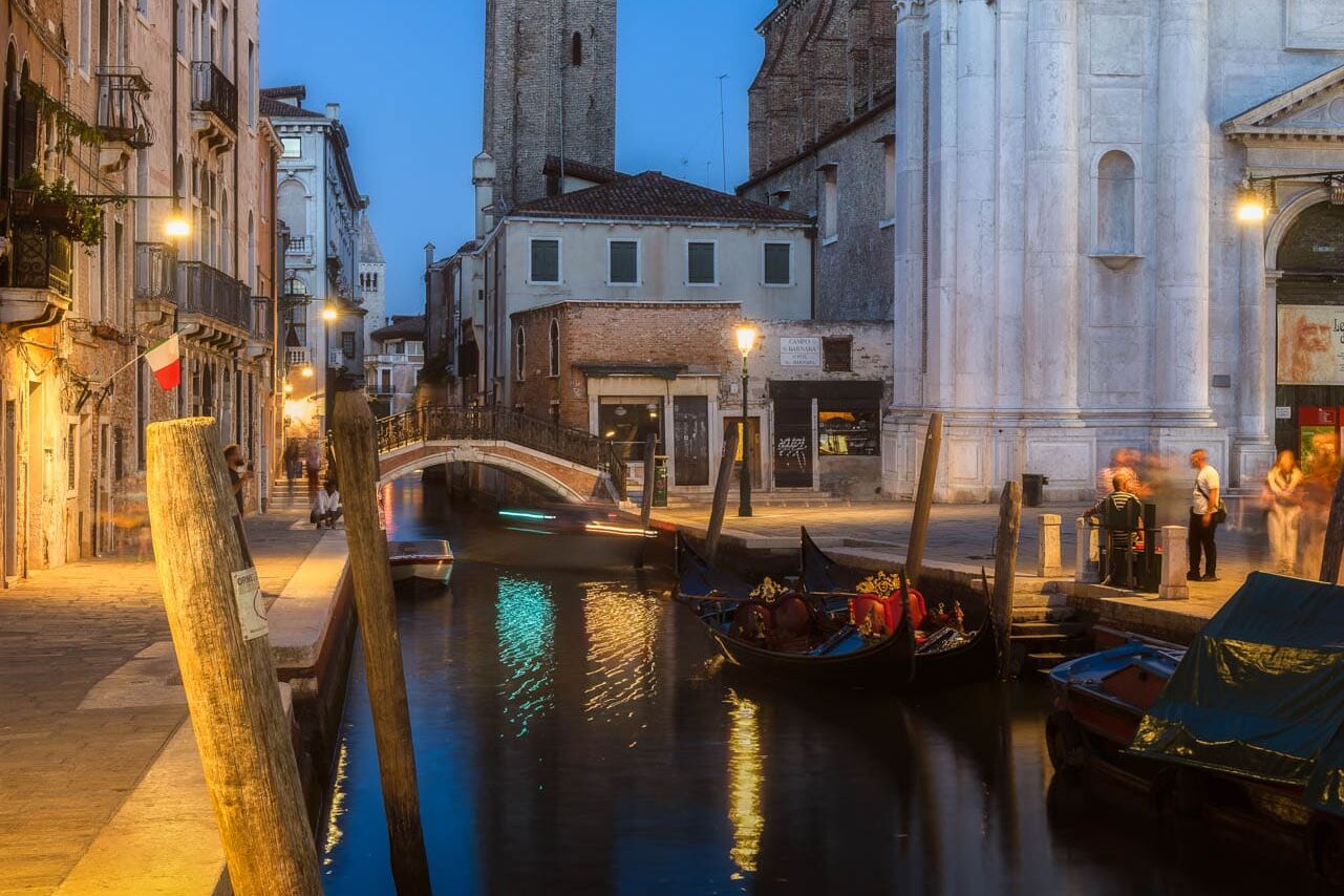 Venetië bij blauw uur: gondels langs een kanaal, pastelgevels, witte marmeren gevel, boogbrug en lampen.