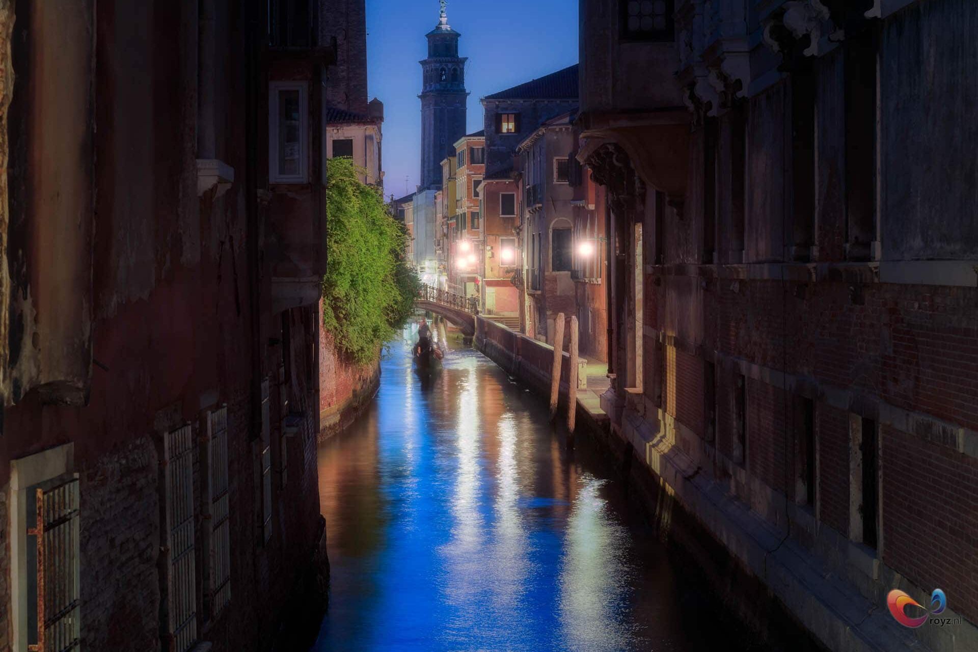 Nachtkanaal in Italiaans stadje met brug, toren en spiegelingen op kalm water.