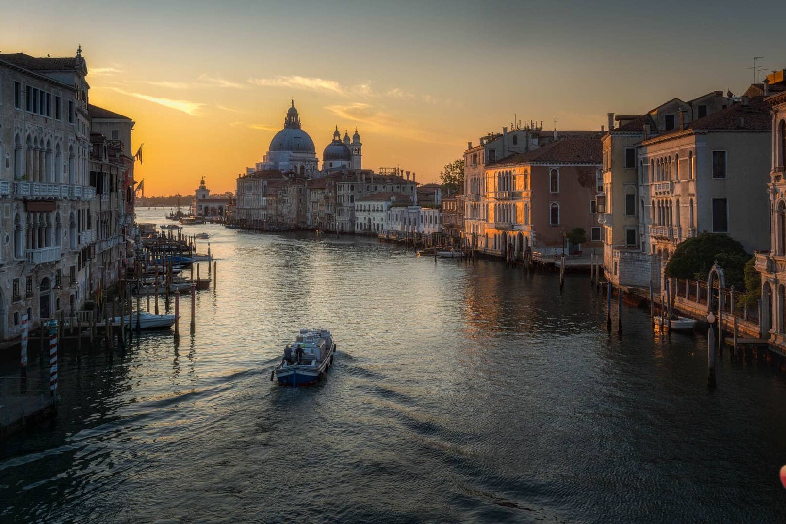 Venetië bij zonsondergang: motorboot over kalm kanaal, palazzi en koepelkerk aan de horizon.