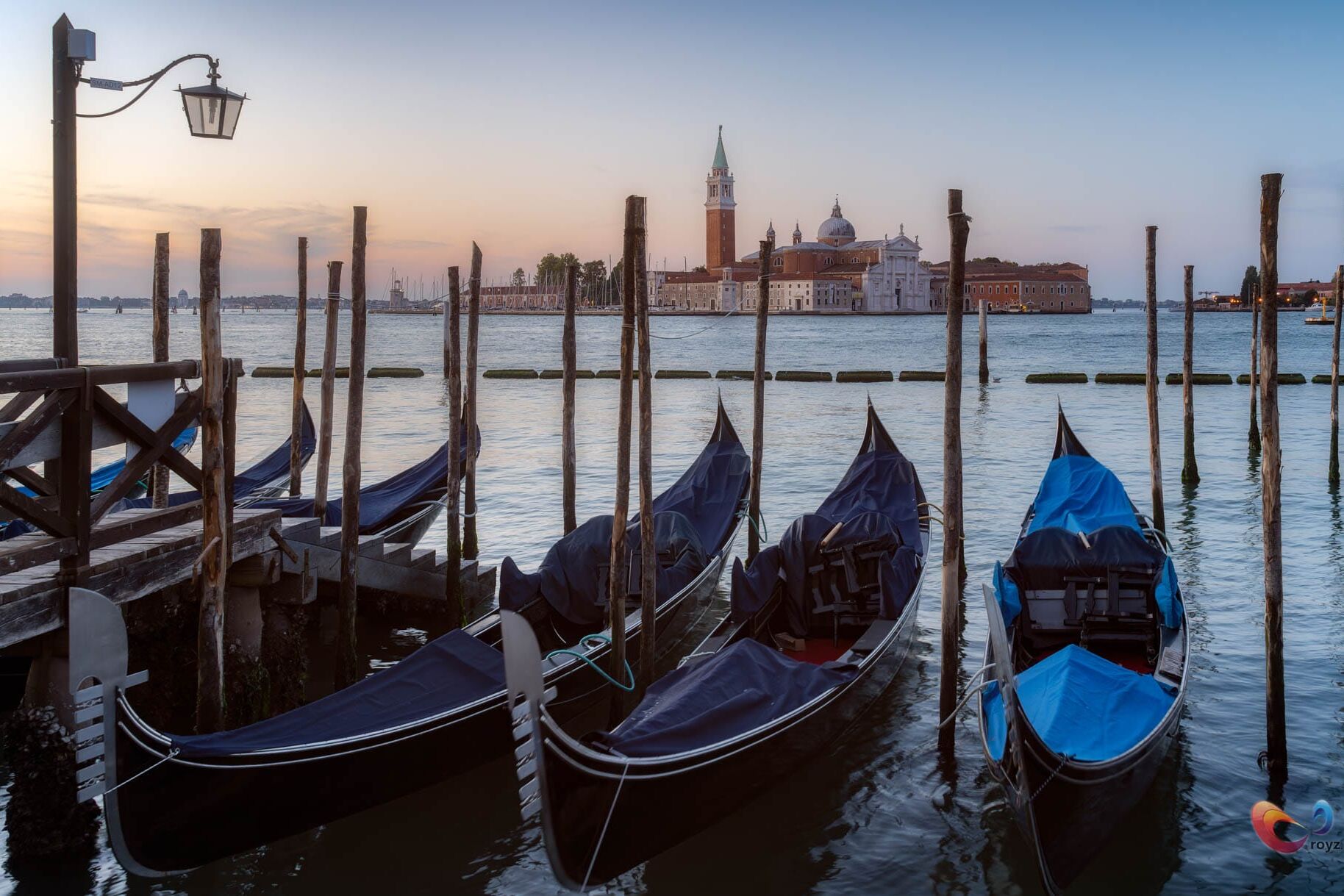 Venetië bij zonsondergang: gondolas, houten dok, toren en witte koepelkerk aan de horizon.