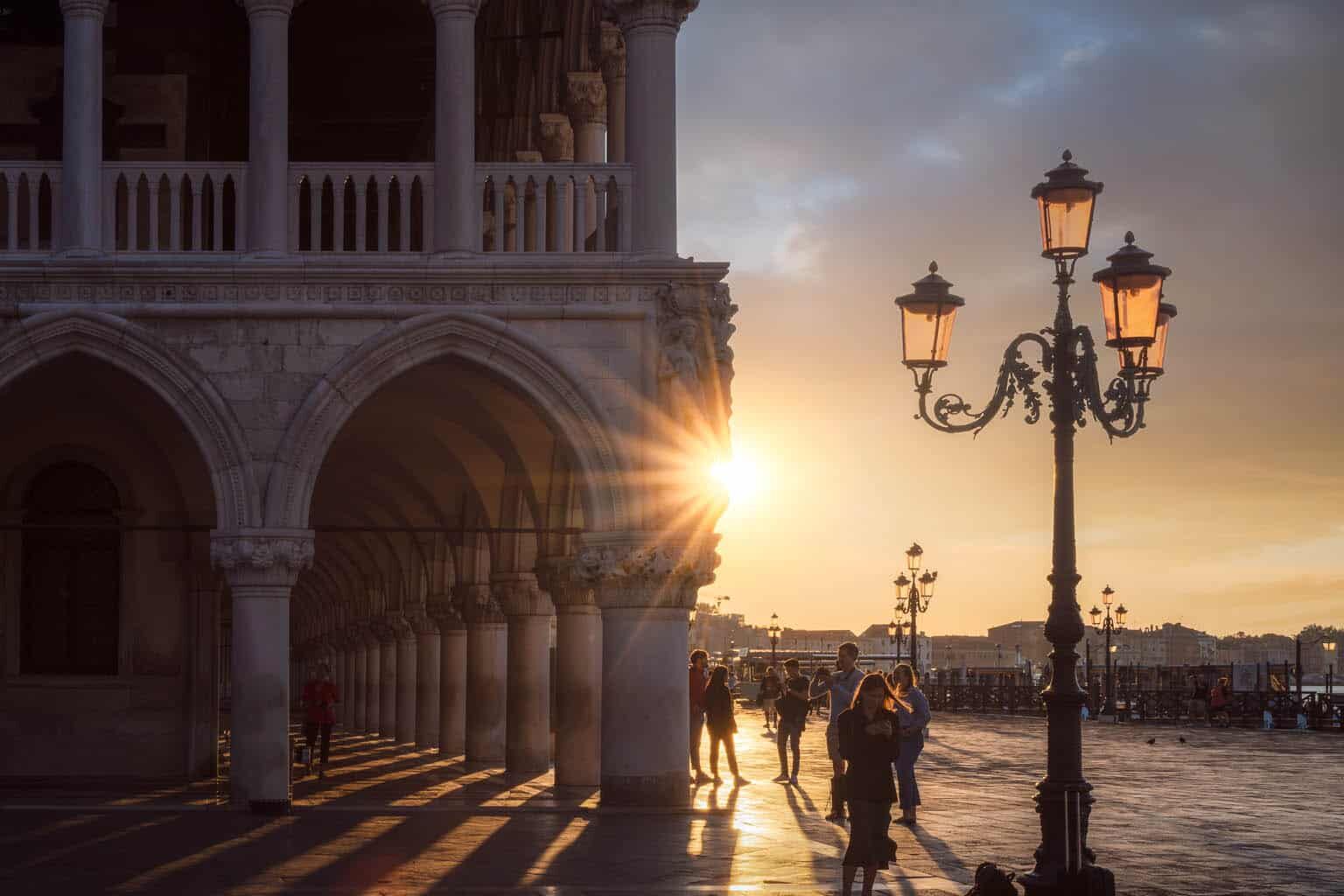 Venetië: gotische marmeren loggia bij zonsondergang, reizigers, straatlantaarns en kasseien plein.