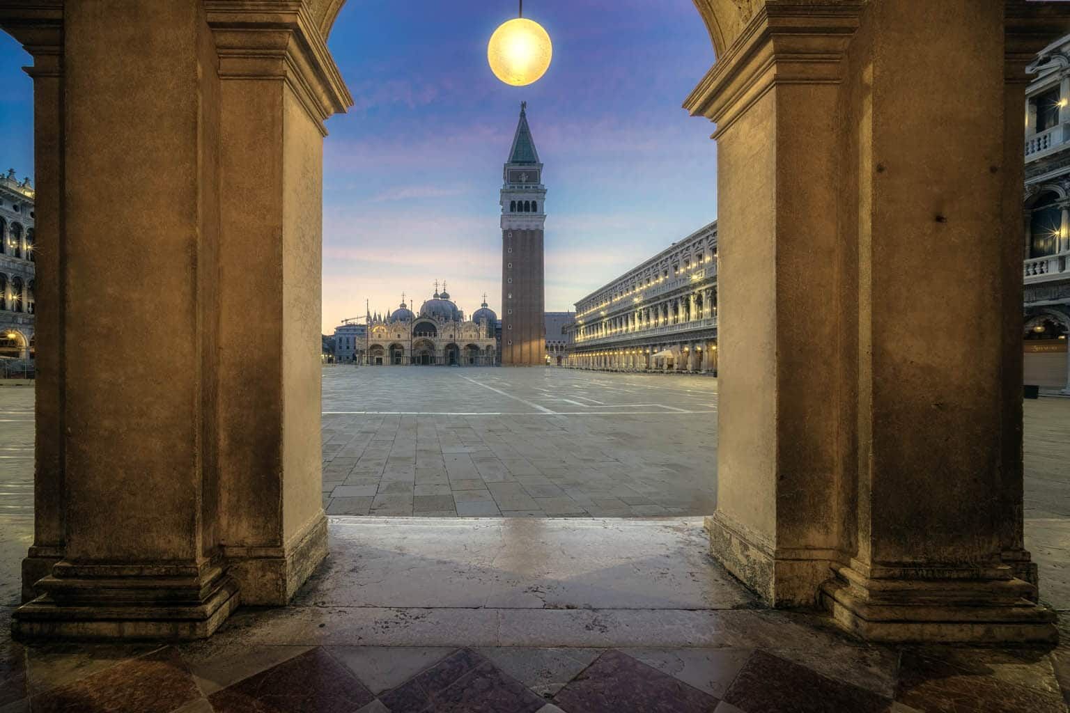 Venetië bij schemering: poort met amber lamplicht, Campanile en San Marco Basiliek boven arcade-piazza.