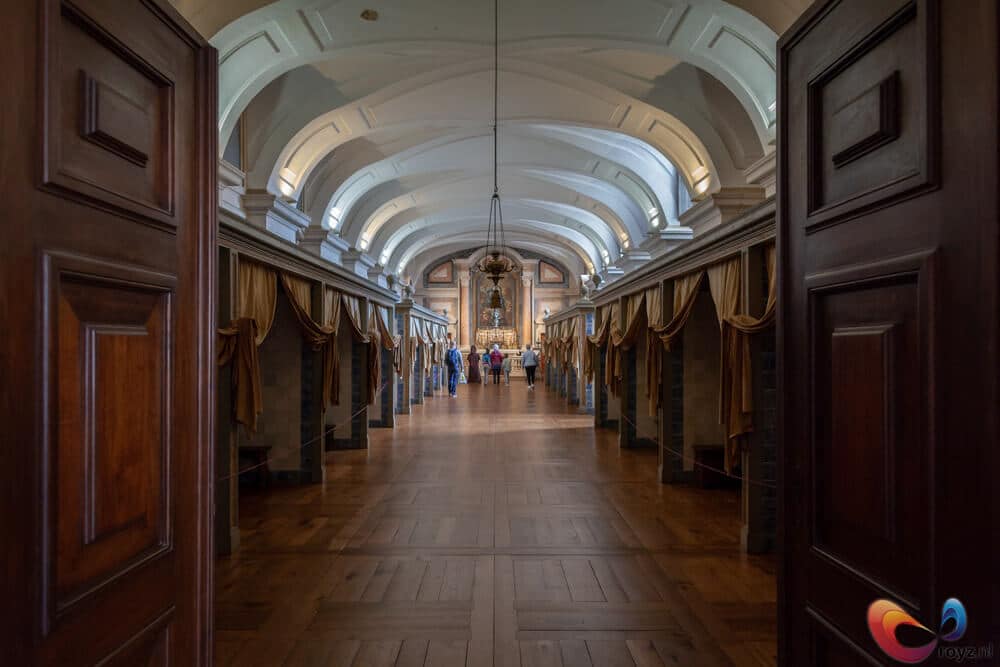Historische gang in paleis met tongewelven, gordijnen, warme houten vloer en kroonluster.