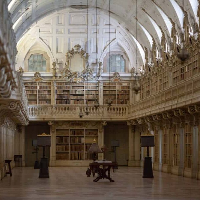 Historische bibliotheekhal met dubbele galerijen, rococo-ornamenten, centrale ronde tafel en warme verlichting.