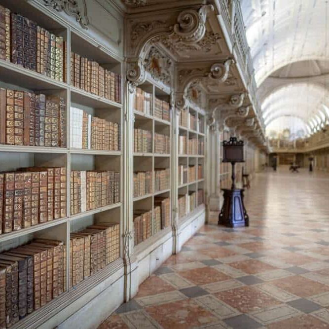 Lange bibliotheekhal met ivoorkleurige boekenkasten, donkere lessenaar en galerij met balustrade onder daglicht.