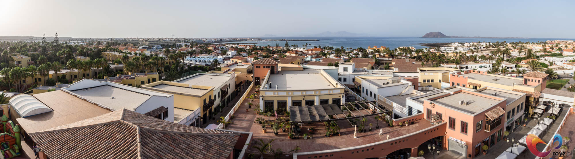 Corralejo: cirkelvormig bakstenen terras naast wit kolomgebouw langs promenade bij kalme zee.